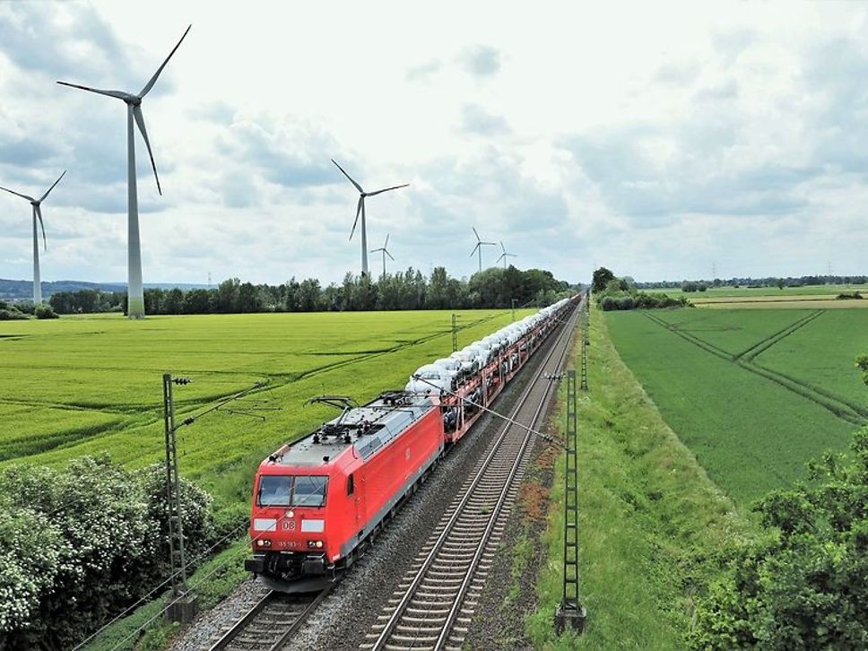 Ein Autotransportwagen fährt durch eine grüne Landschaft, im Hintergrund sind Windmühlen zu sehen. 